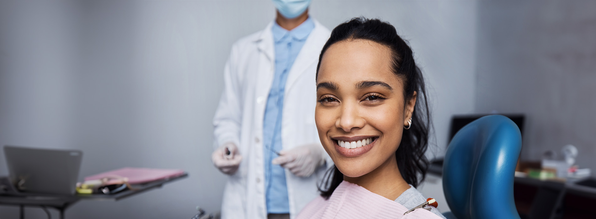 A smiling dental hygienist in a professional setting, wearing a face mask and blue scrubs, standing behind a patient chair with a patient seated.