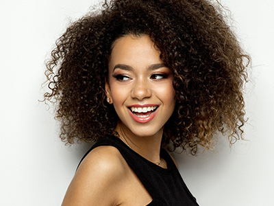 The image features a woman with curly hair, wearing makeup and a black top, smiling and posing for the camera.