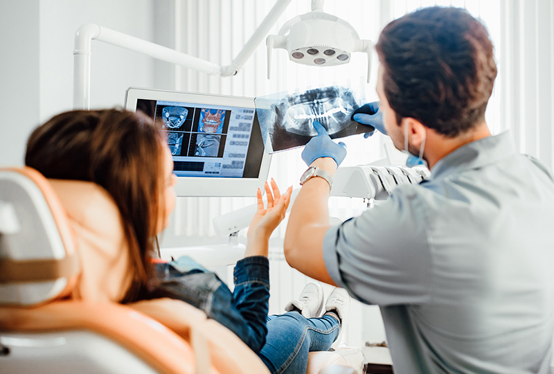 A dental professional is assisting a seated patient with a dental chair, using a digital display to show the patient s x-ray.
