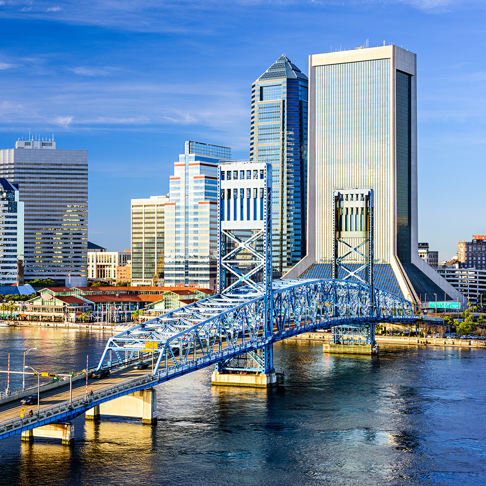 A vibrant cityscape with a prominent bridge, skyscrapers, and boats on the river under a clear blue sky.